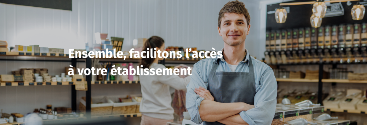 Une photo horizontale en plan moyen du même jeune homme souriant en tablier de magasin, bras croisés, mais dans un plan plus large montrant l'épicerie en vrac plus en détail. Le magasin dispose d'un comptoir central avec une caisse enregistreuse, de plus d'étagères de produits en vrac, et d'un point de vente flou au premier plan à gauche. Le texte blanc et gras "Ensemble, facilitons l'accès à votre établissement" est superposé sur la gauche. Une femme floue se tient à l'arrière-plan près des étagères.