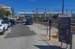 vue de la zone dépose-minute du parking EFFIA de la Gare de Béziers : le panneau d'information au premier plan détaille les tarifs et services, tandis que l'on aperçoit la passerelle de la gare et l'accès sécurisé pour les voyageurs en arrière-plan.