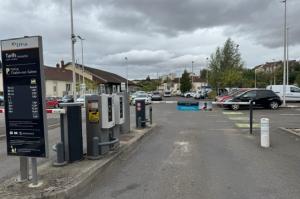 Voie d'accès au parking extérieure équipée de bornes de contrôle à tickets, d'une barrière levante et d'un panneau d'affichage des tarifs.
