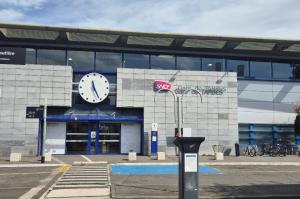 Façade de la gare de Tarbes avec son horloge centrale, l'accès à la gare routière, un horodateur et des emplacements réservés au premier plan.