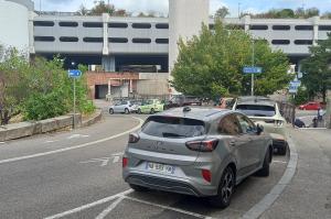 Voie de circulation en courbe longeant un parking, avec véhicules stationnés et structure en béton d'un parking silo en arrière-plan.