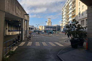 Perspective depuis un passage couvert donnant sur une rue extérieure avec des immeubles et des bacs de fleurs.