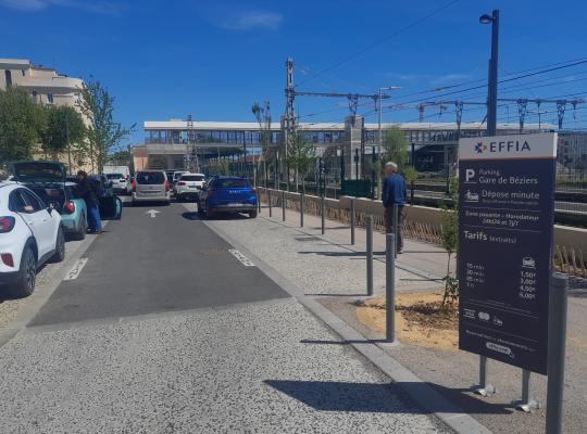 vue de la zone dépose-minute du parking EFFIA de la Gare de Béziers : le panneau d'information au premier plan détaille les tarifs et services, tandis que l'on aperçoit la passerelle de la gare et l'accès sécurisé pour les voyageurs en arrière-plan.
