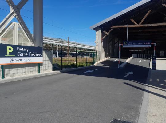 vue de l'accès extérieur au parking de la Gare de Béziers géré par EFFIA, avec un grand panneau de bienvenue "24/24" : l'image capture la rampe d'entrée menant au bâtiment couvert à charpente bois, juxtaposée aux voies ferrées et à l'infrastructure de la gare.