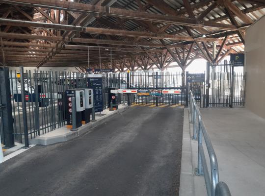 découvrez l'entrée sécurisée du parking EFFIA couvert de la Gare de Béziers, sous une impressionnante charpente en bois : le visuel montre les barrières automatiques, les bornes de péage et la signalétique claire pour le stationnement des voitures et des vélos.