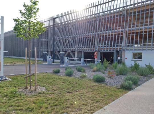 Façade en bois moderne et accès au parking EFFIA Valmy à Dijon : voies d'entrée sécurisées avec barrières automatiques, bornes de ticket et aménagement paysager soigné à proximité.