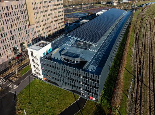 Vue aérienne du parking EFFIA de la gare de Caen : un ouvrage moderne avec toiture photovoltaïque, situé entre les voies ferrées et de nouveaux immeubles de bureaux.