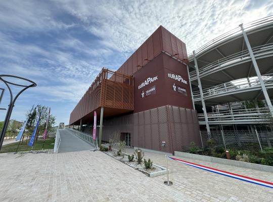 Façade moderne du parking EuraPark à Douai : un ouvrage aérien avec habillage métallique brun, rampe d'accès piétonne et parvis aménagé.