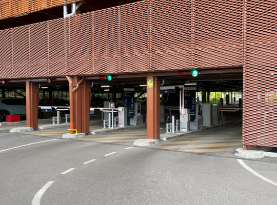 Accès principal au parking couvert EFFIA : l'image montre les voies d'entrée et de sortie sécurisées, équipées de barrières automatiques, de bornes de péage et de feux de signalisation sous une façade moderne en résille métallique teinté terre cuite.