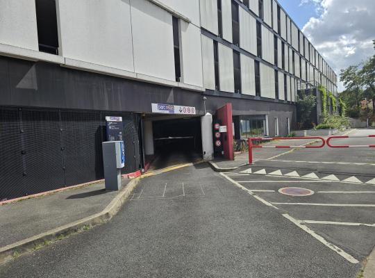 Voie d'accès au parking couvert avec borne de péage et barrière automatique.