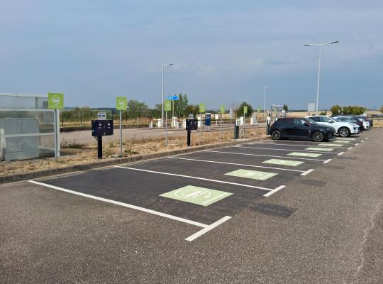 Alignement de places de parking asphaltées réservées aux véhicules électriques, signalées par des marquages au sol verts et des bornes de recharge à écran.