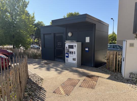 Caisse automatique de paiement installée contre un petit bâtiment gris foncé.