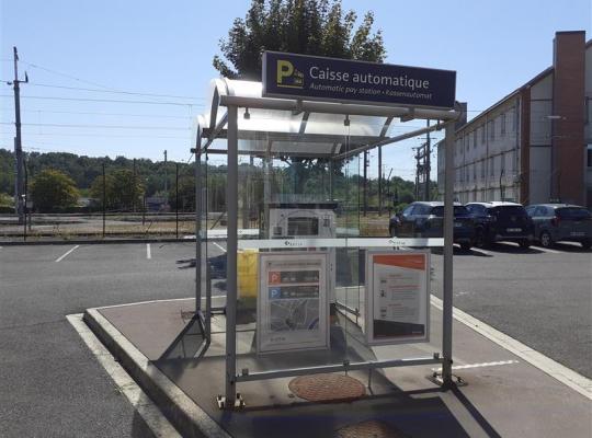 Caisse automatique de paiement protégée sous une structure vitrée couverte sur le parking.