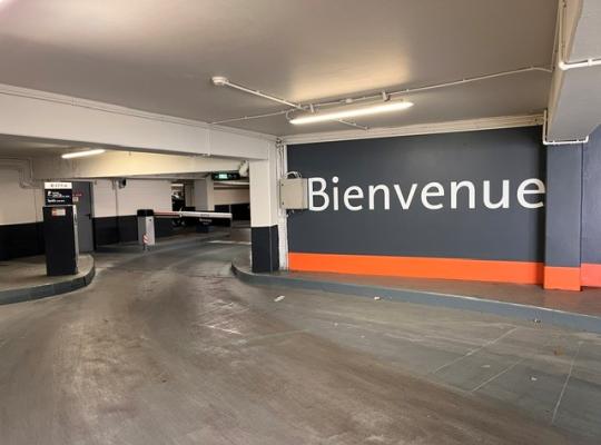 Voie d'accès couverte du parking avec borne d'entrée, barrière automatique et mur d'accueil signalétique.