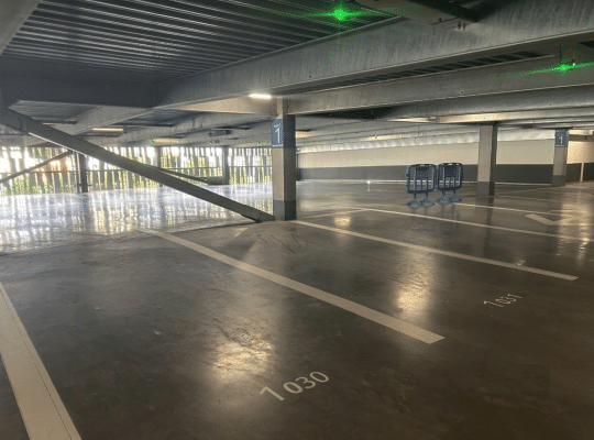 Allée de stationnement au niveau 1 d'un parking couvert souterrain à structure métallique moderne. Le sol en béton lisse comporte des marquages de places numérotés et un poteau porte la signalétique bleue du niveau 1.