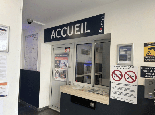Bureau d'accueil EFFIA dans un hall de parking bien éclairé. Une signalétique "ACCUEIL" noire et blanche claire surplombe la zone de service. Un panneau de sécurité "PARKING SOUS ALARME ET VIDÉOPROTECTION" jaune et un plan d'intervention sont visibles sur le mur.