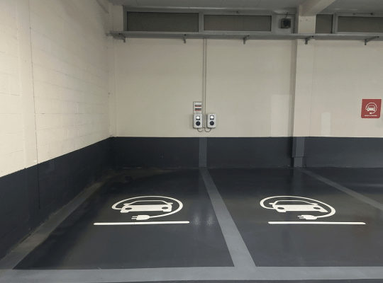 Vue rapprochée de deux places de stationnement réservées aux véhicules électriques dans un parking souterrain. Un logo blanc distinctif représentant une voiture en charge est peint au sol. Un panneau mural rouge de signalisation est également visible.