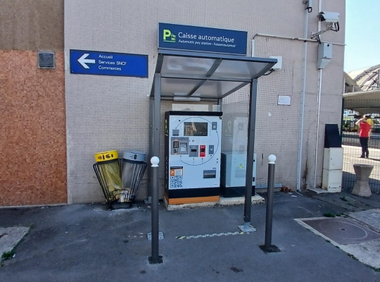 Caisse automatique de paiement installée sous un abri vitré contre le bâtiment.