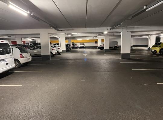 Intérieur du parking couvert éclairé avec places de stationnement délimitées et piliers structurants.
