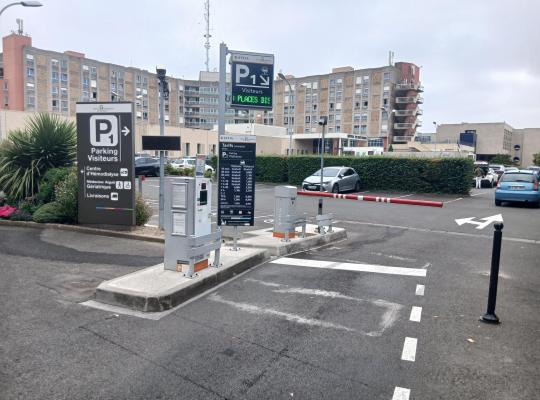 Borne de contrôle d'accès, barrière automatique et panneaux d'information à l'entrée du parking P1 de l'hôpital.