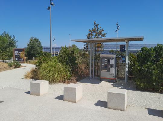 Caisse automatique EFFIA sous un abri vitré, bordée de blocs de béton et de végétation sur le parking.