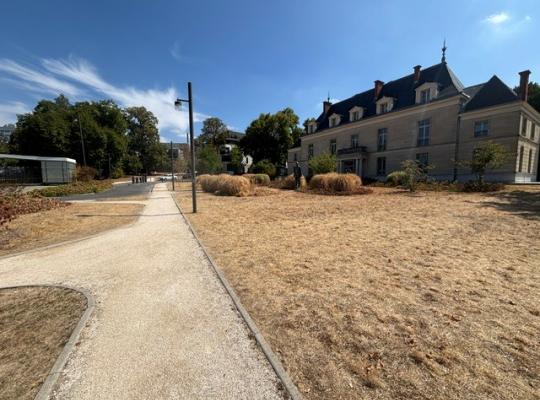 Chemin piéton menant à une grande bâtisse en pierre au bord du parking.