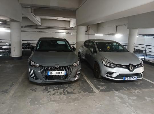 Une vue intérieure d'un parking souterrain éclairé par des tubes fluorescents, avec des piliers en béton. Deux voitures, une Peugeot grise au premier plan et une Renault argentée derrière, sont garées en marche avant dans des places marquées au sol par des lignes jaunes. Le plafond est bas, révélant des conduits et des tuyaux. Des grilles de fer noir séparent les allées. Des panneaux d'information ou des numéros de place sont visibles sur les piliers ou les murs à l'arrière.