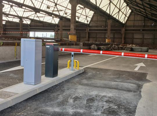 Voie d'accès couverte sous une structure métallique, équipée d'une borne de paiement et d'une barrière automatique rouge et blanche.