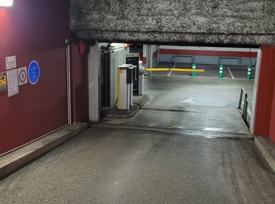 Rampe d'accès intérieure bordée de murs rouges menant aux bornes et barrières du parking.