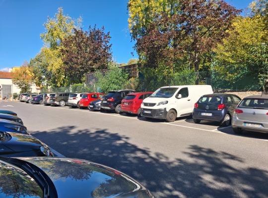 Alignement de véhicules stationnés sur le parking extérieur, longé par un espace vert et des arbres.