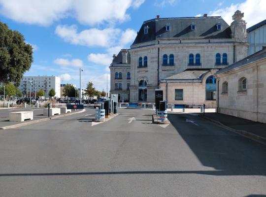 Accès au parking avec barrières automatiques et bornes de paiement devant le bâtiment historique de la gare.