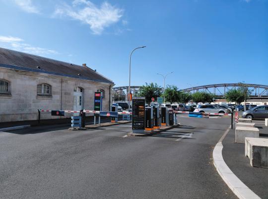 Voies d'entrée et de sortie du parking extérieures, équipées de bornes de contrôle à tickets, de barrières et d'un panneau tarifaire.