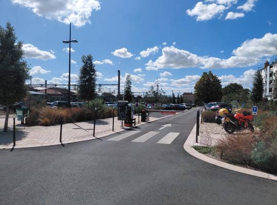 Voie d'accès extérieure au parking équipée d'une borne d'entrée à ticket, d'une barrière levante et d'un panneau d'affichage des tarifs.