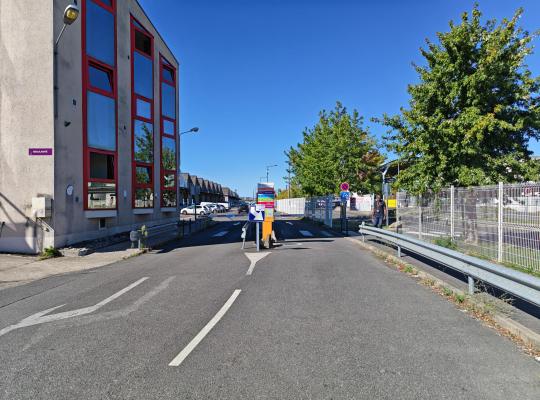 Entrée du parking extérieur longeant un bâtiment vitré aux cadres rouges.