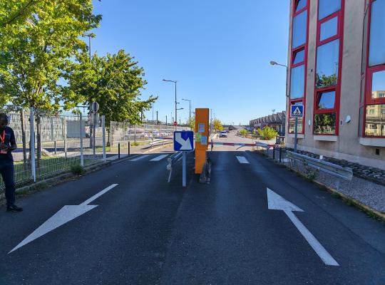 Voies d'accès et de sortie du parking équipées d'une borne orange et d'une barrière automatique.