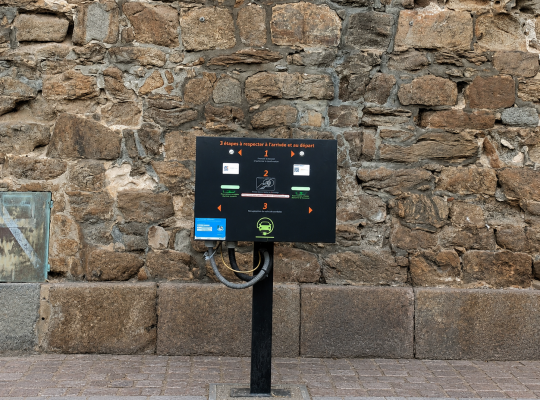 Borne de recharge pour véhicules électriques fixée au sol devant la muraille en pierre du rempart.