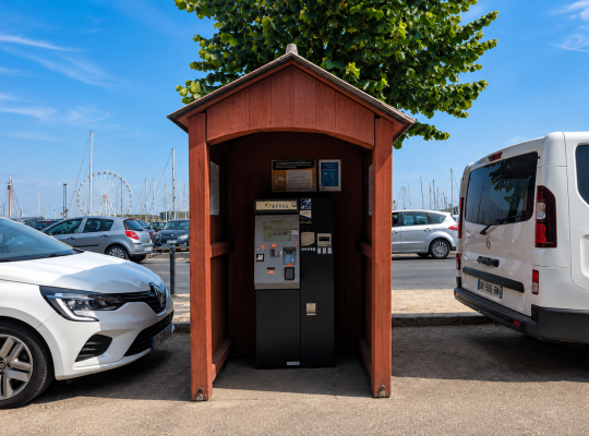 Borne de paiement automatique installée à l'abri sous un petit chalet en bois rouge entre les places de stationnement.