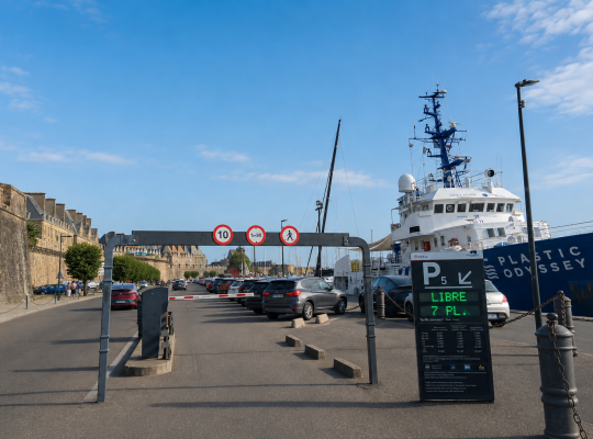 Entrée du parking P5 avec portique de hauteur, panneau d'affichage des places et navire amarré à quai.