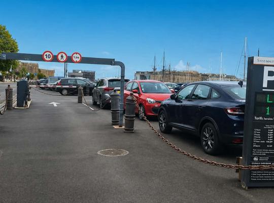 Entrée du parking avec portique de gabarit, barrière d'accès et panneau d'affichage des tarifs et places disponibles.