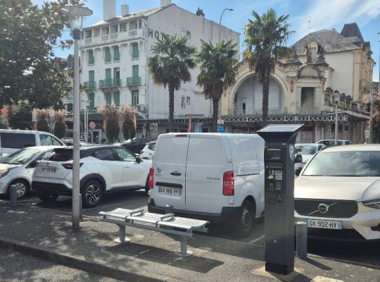 Zone de stationnement en épi le long de la place de la Gare, avec horodateur, banc public et véhicules alignés devant des bâtiments anciens et palmiers.