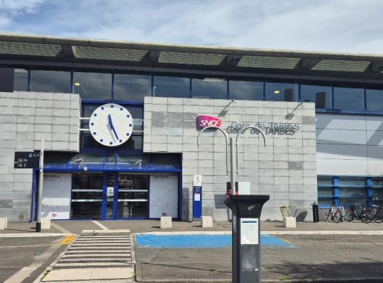 Façade de la gare de Tarbes avec son horloge centrale, l'accès à la gare routière, un horodateur et des emplacements réservés au premier plan.