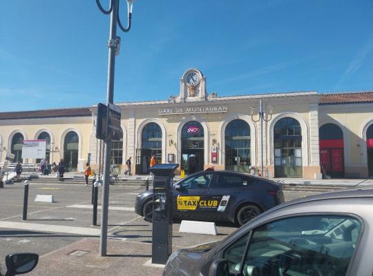 Façade historique de la gare de Montauban-Ville-Bourbon avec son horloge centrale, un horodateur et un taxi garé sur le parvis.