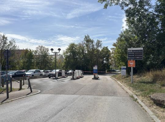 Voie d'accès au parking de l'hôpital bordée d'arbres, équipée de barrières automatiques et d'un panneau d'information EFFIA.