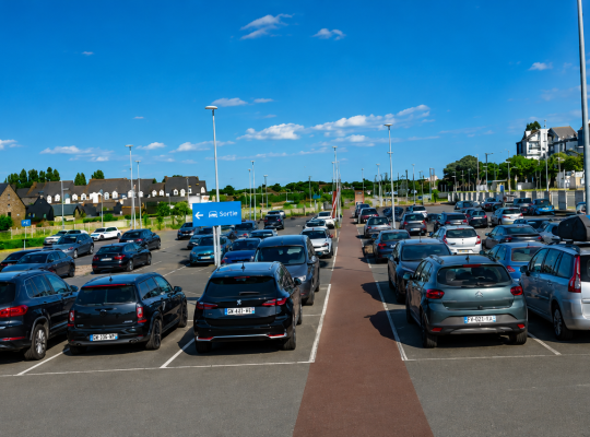 Grand parc de stationnement extérieur avec de nombreuses places balisées à proximité immédiate des voies ferrées et des logements.