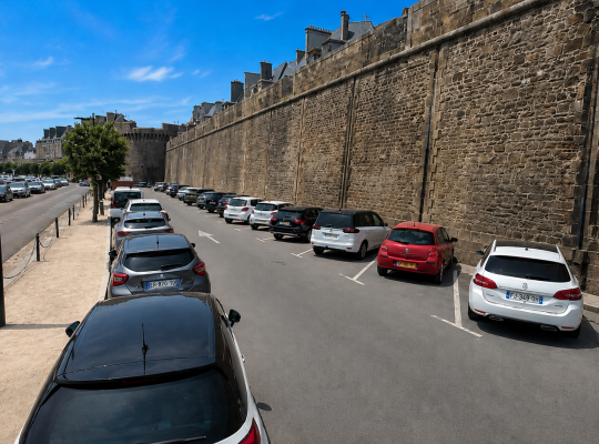 Rangée de véhicules stationnés le long de la muraille historique, en bordure de la voie de circulation.