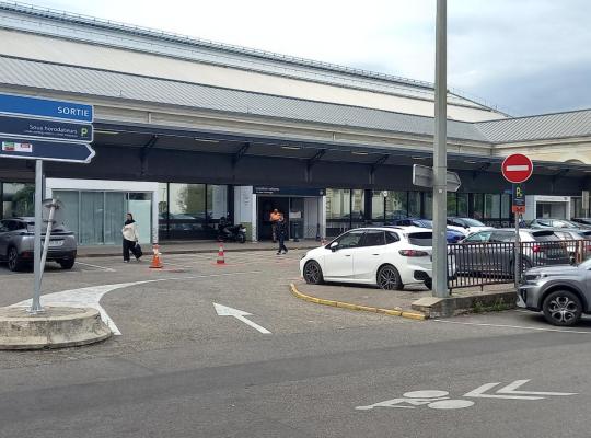 Parvis avec panneaux de signalisation de sortie et de parking, zone de dépose-minute et abri pour voyageurs devant la gare.