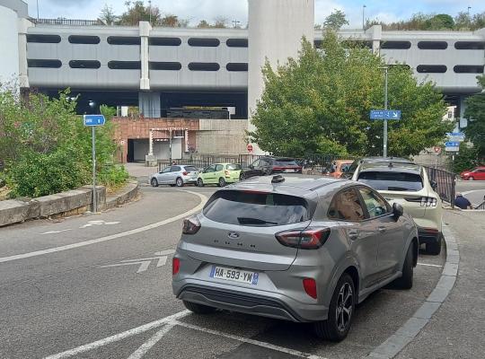 Voie de circulation en courbe longeant un parking, avec véhicules stationnés et structure en béton d'un parking silo en arrière-plan.