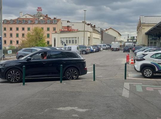 Aire de stationnement avec véhicules en autopartage et passage piéton, située devant la halle et les bâtiments environnants.