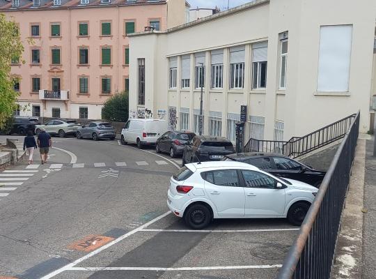 Voie en descente bordée de places de stationnement en épi le long d'un bâtiment administratif, avec piétons sur la chaussée.