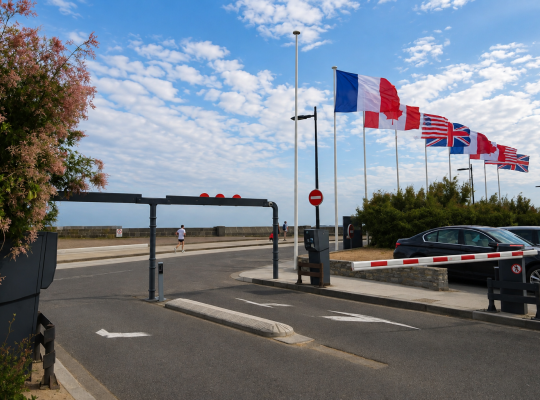 Voie de sortie du parking avec barrière automatique, bordée par une rangée de drapeaux internationaux.